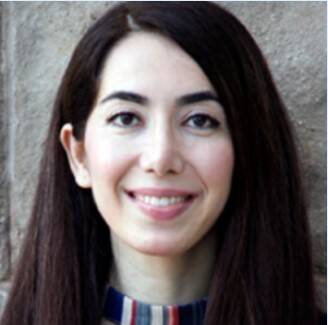 A person with long, dark hair smiles at the camera. They are wearing a colorful, striped necklace and a neutral top. The background is a textured, stone wall.
