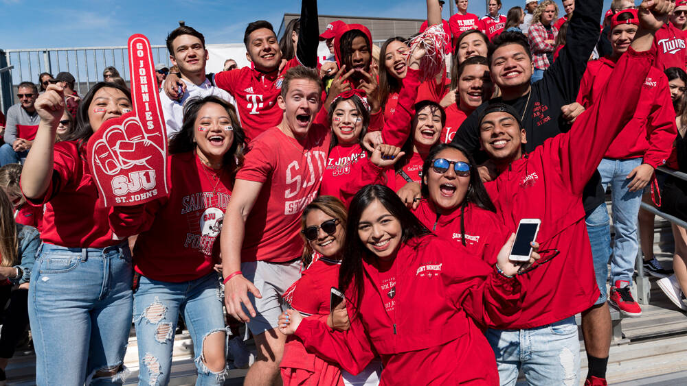 A large group of enthusiastic people, all dressed in red, gather closely together outdoors, smiling and cheering. They hold up various signs and foam fingers with the words "Go Johnnies" and "SJU" visible. The sky is clear and blue in the background.