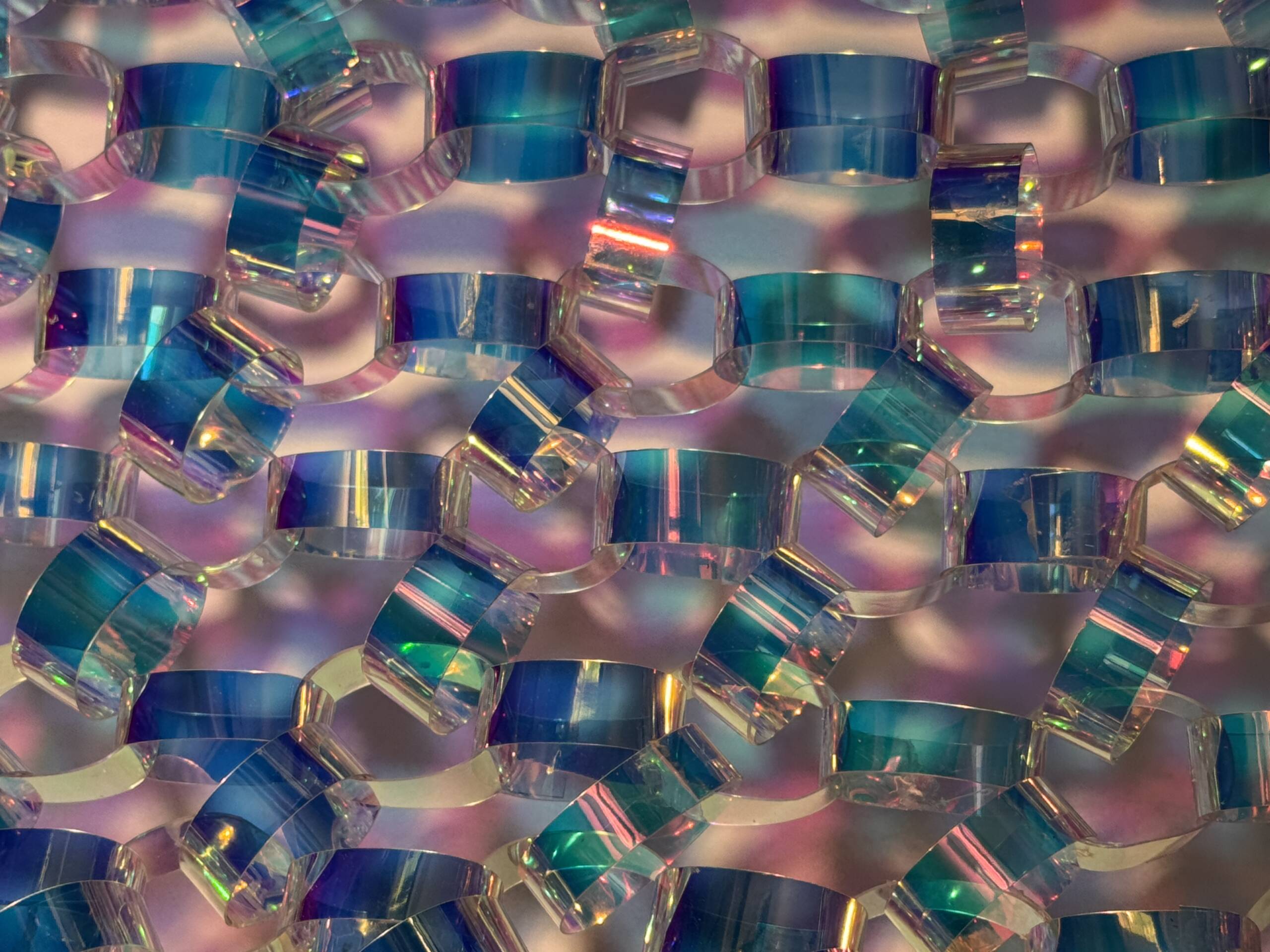 A close-up of overlapping translucent plastic or glass links with iridescent blue, green, and purple hues, creating a vibrant, abstract pattern against a soft, multicolored background.