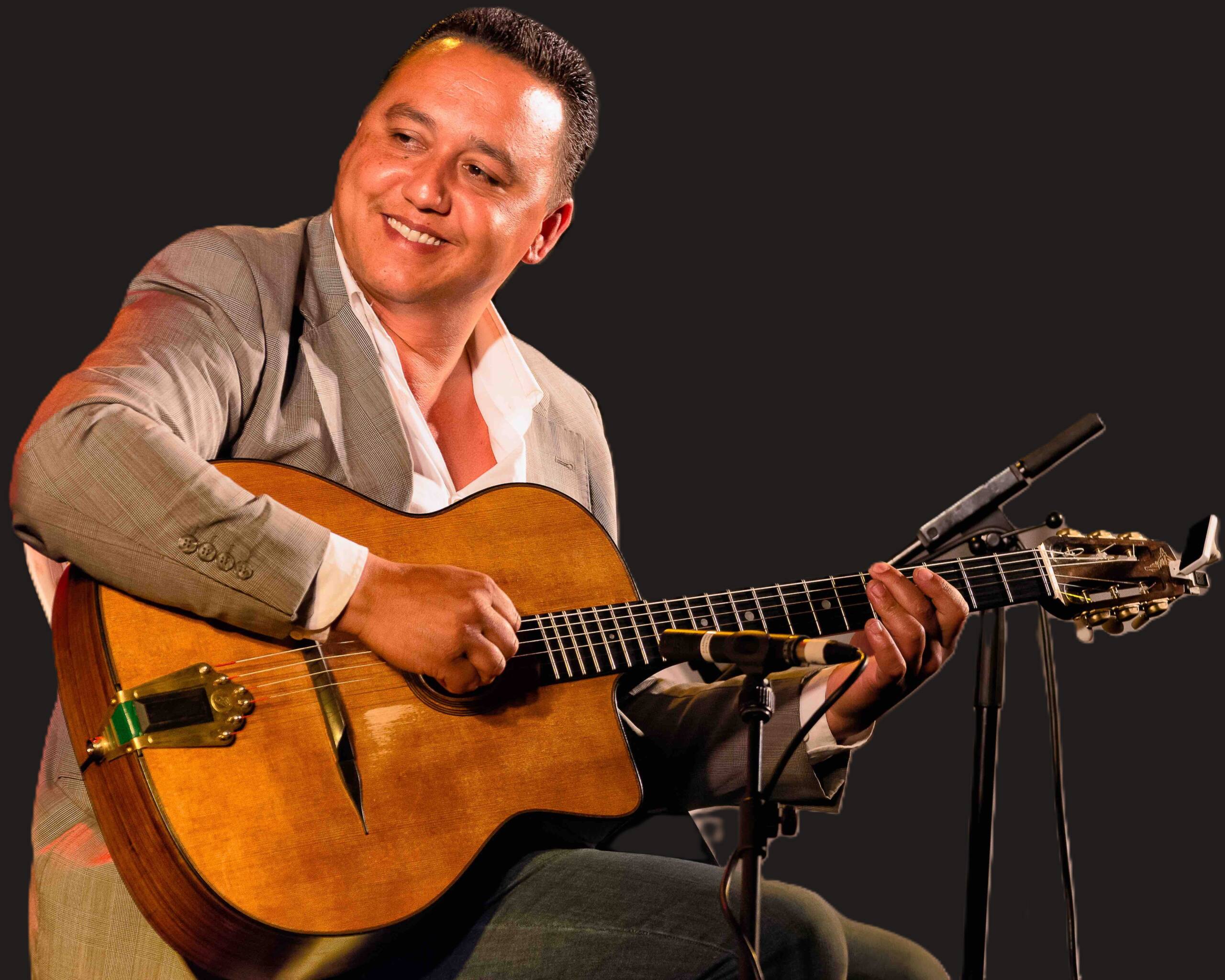 A man in a grey suit smiles while playing an acoustic guitar. He is seated and surrounded by microphones against a dark background.