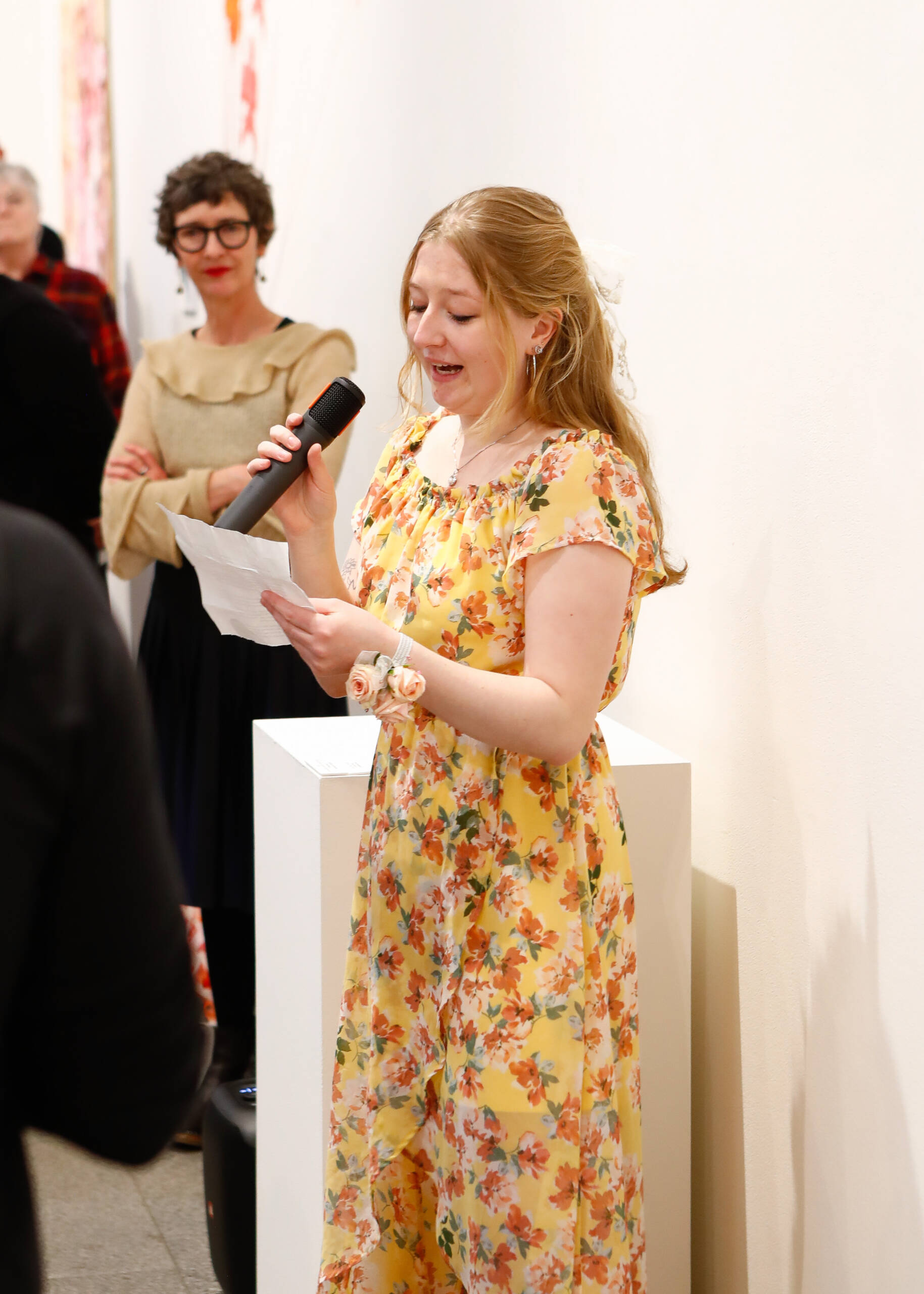 A young woman in a yellow floral dress speaks into a microphone while holding a piece of paper at an indoor event. She is smiling. In the background, another woman stands and listens.