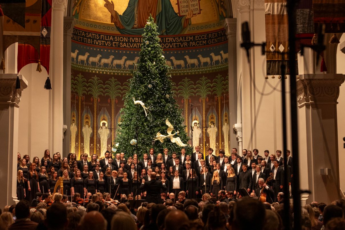 A large choir performs in a grand church decorated for Christmas, with a tall, lit Christmas tree and doves in front of a mural. An audience is seated in front, watching the performance.