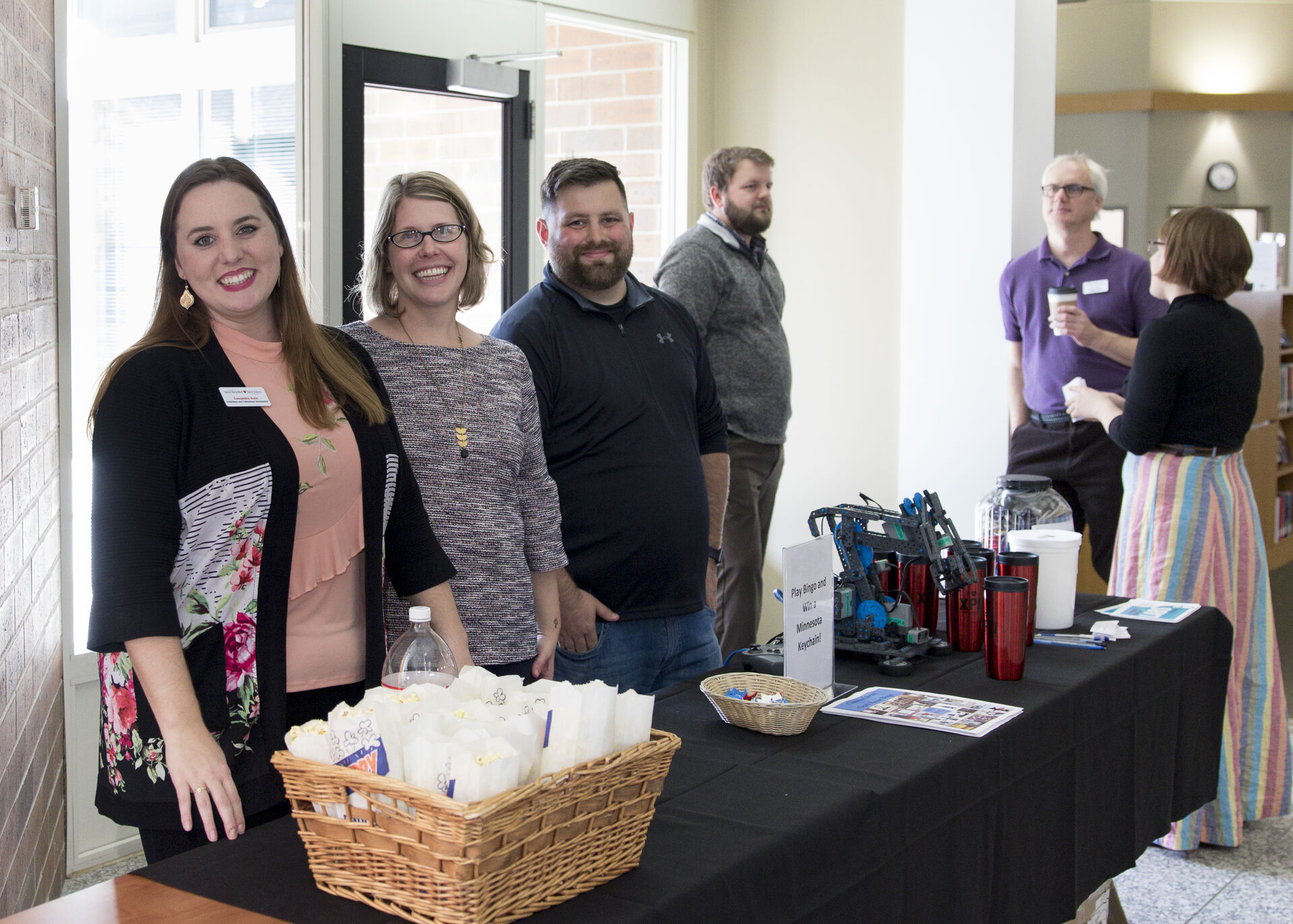 Five people stand behind a table with snacks, drinks, and promotional items at a well-lit indoor event; one woman in a floral jacket smiles at the camera while others interact with attendees.