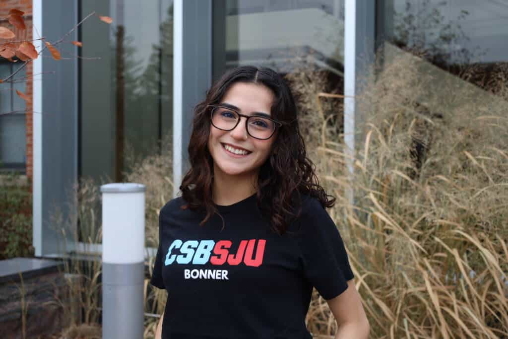 A young woman with wavy brown hair and glasses smiles outside, wearing a black "CSB SJU Bonner" t-shirt. She stands in front of tall grasses and modern windows.