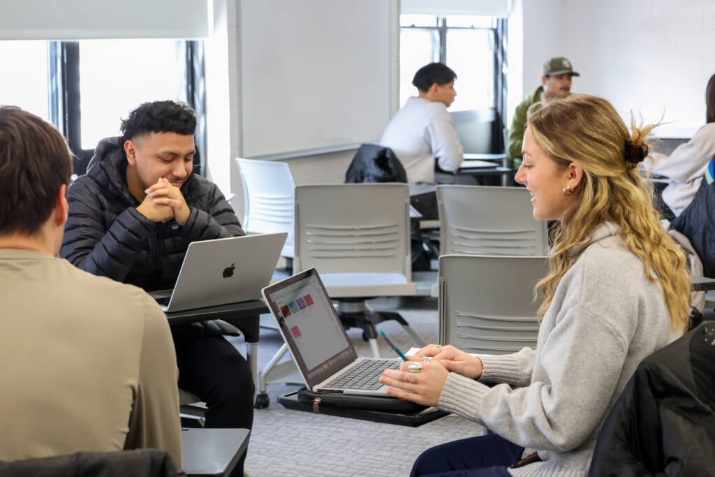 Three students sit at desks in a classroom, two of them facing each other and smiling, with laptops open. Other students are visible in the background, also working and talking in groups.