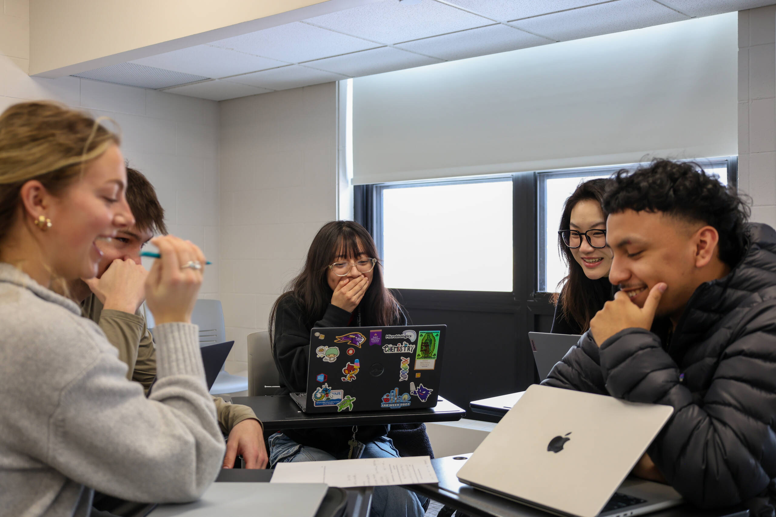 Five students sit at desks in a classroom, smiling and laughing together. Laptops with stickers are visible, and sunlight streams through the windows behind them, creating a bright and cheerful atmosphere.