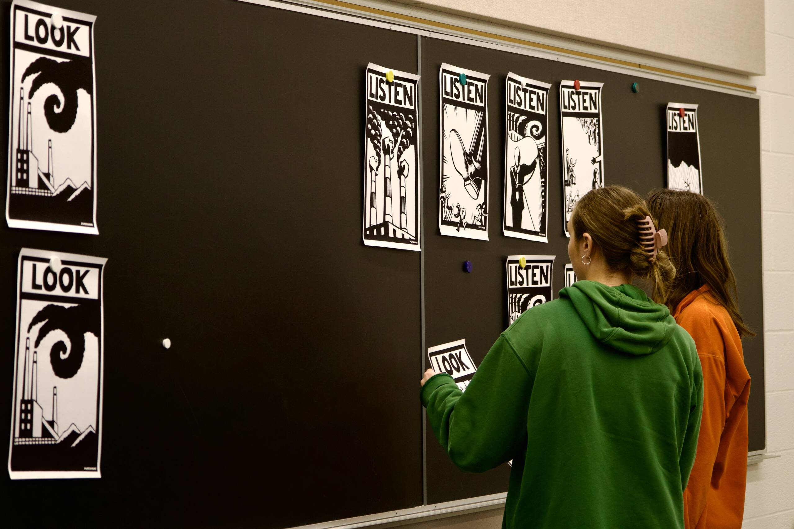 Two people stand in front of a blackboard with posters that say "LOOK" and "LISTEN," featuring black-and-white graphic art. One person holds a "LOOK" poster, while the other examines the display.