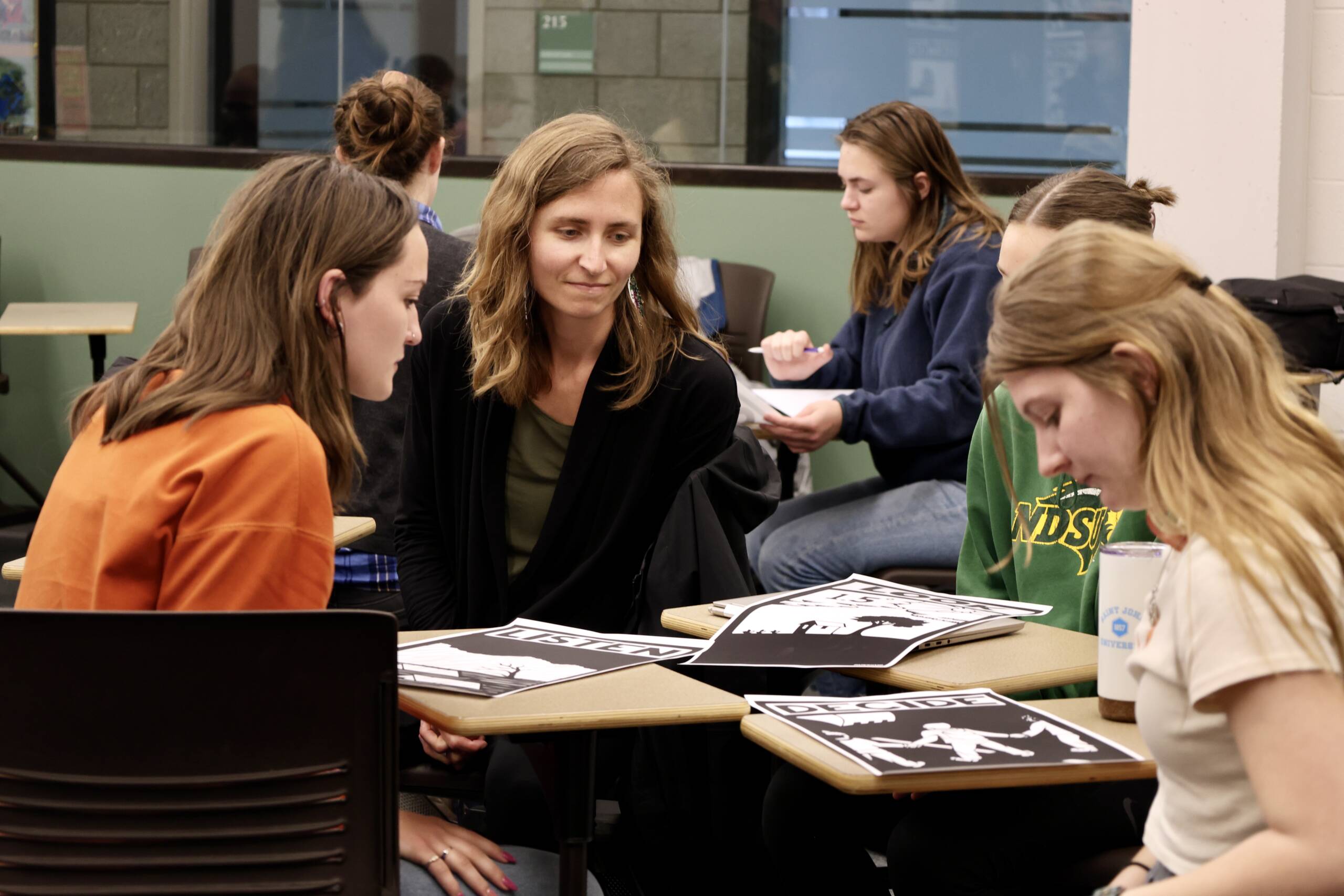 Four women sit around a table in a classroom, looking at black and white art prints. One woman in a black sweater smiles, engaging with the group. Other students work in the background.