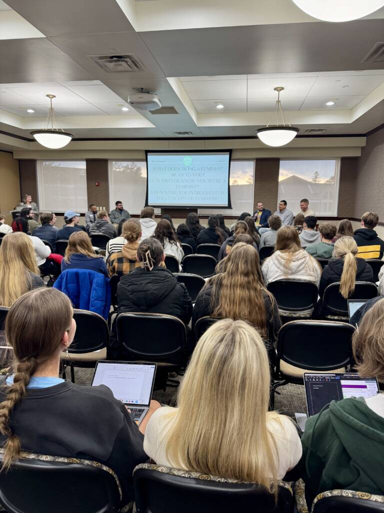 A group of students sit in rows facing a panel of speakers at the front of a conference room, with a large screen displaying text behind the panelists. Some attendees use laptops.