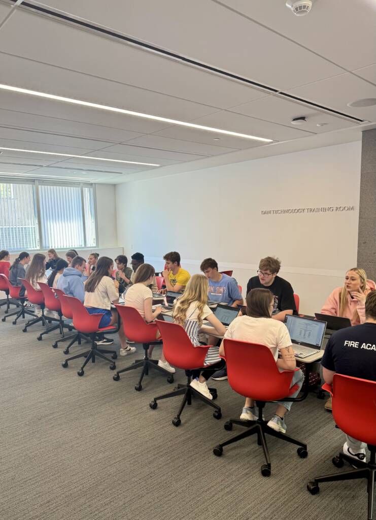 A group of students sits in two long rows facing each other at tables in a bright classroom, working on laptops and talking. They sit in red chairs, and sunlight streams through large windows.