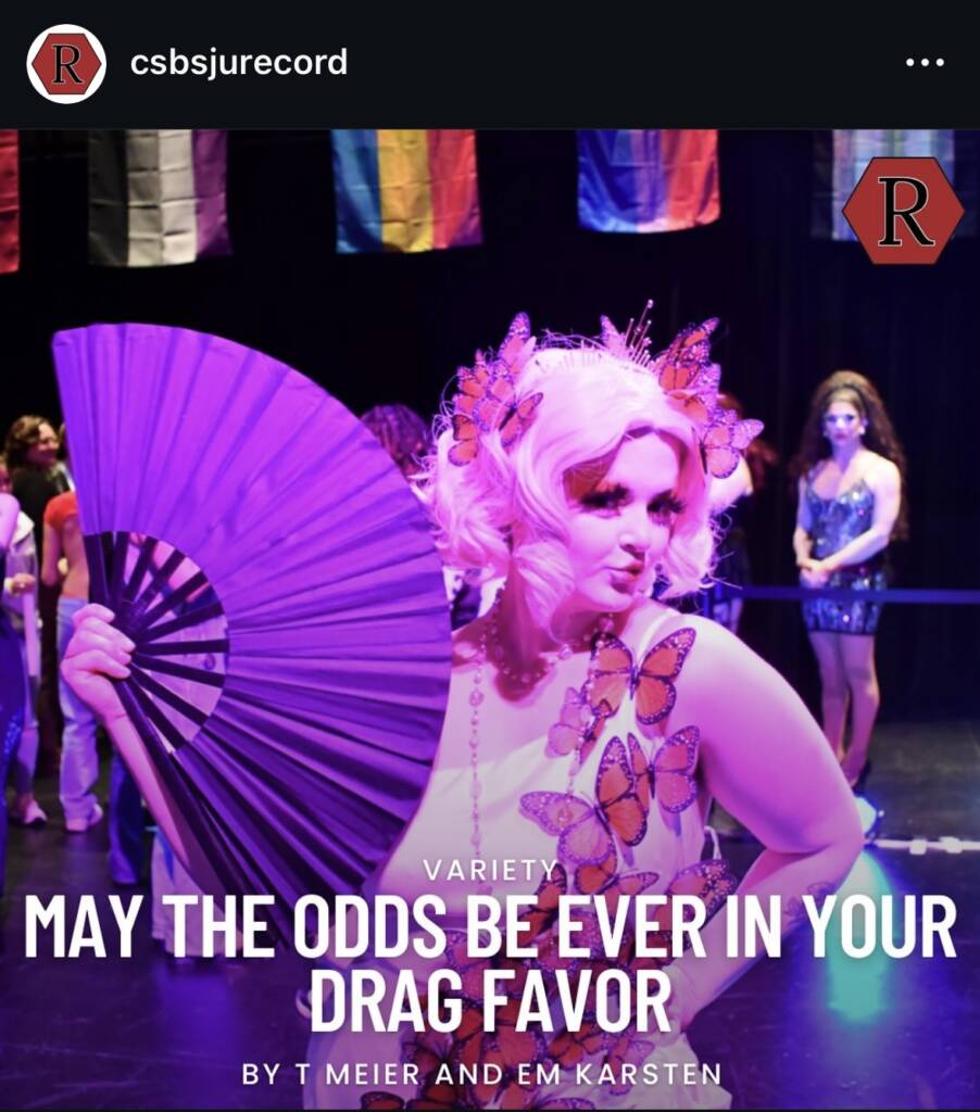 A performer with pink hair, a pink fan, and butterfly decorations poses confidently on stage. Other people and international flags are visible in the background. Text reads: "May the odds be ever in your drag favor.