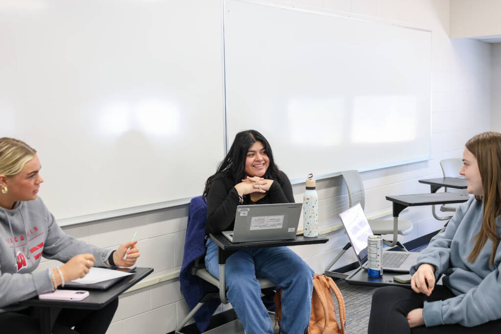 Three students are sitting at desks in a classroom, talking and smiling. Each has a laptop, and there is a whiteboard in the background. One student has a water bottle on the desk.