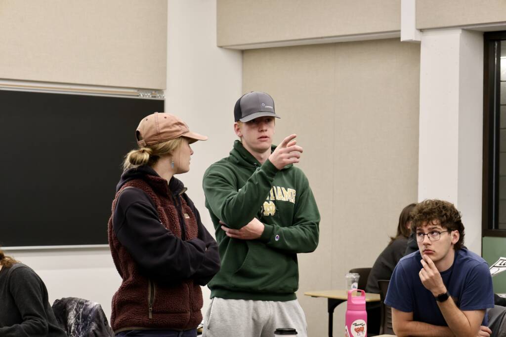 Two students in casual clothes and hats are talking in a classroom; one gestures with his hand. Another student sits nearby, looking thoughtful, with a pink water bottle on the table in front of him.