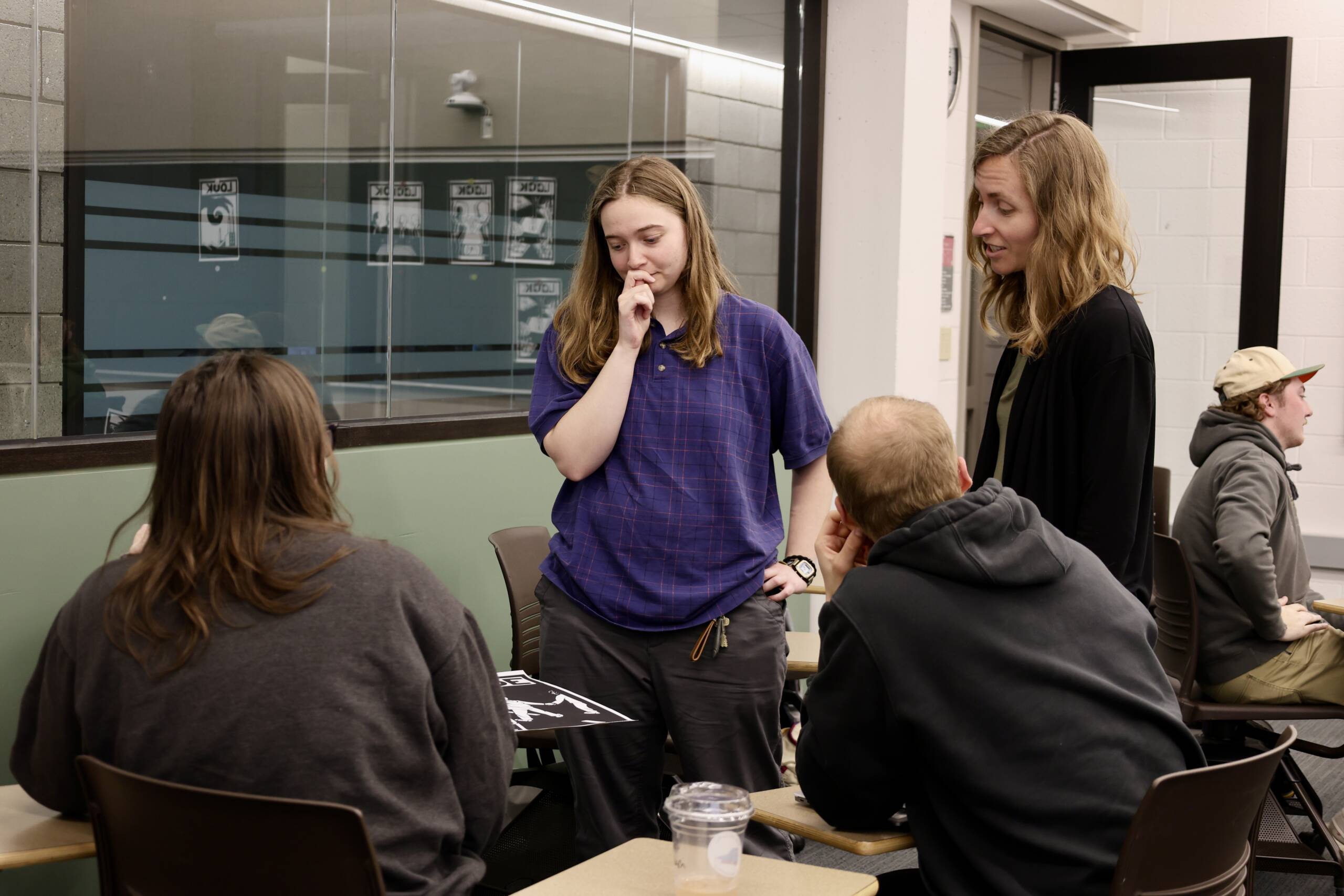 Four people are gathered around a desk in a classroom, engaged in discussion. One woman stands thoughtfully with a hand on her chin, while another stands nearby, speaking to two seated individuals. Another person sits in the background.