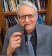 An older person with gray hair is wearing glasses, a gray suit, and a light gray tie. They are holding a pipe and sitting in front of a bookshelf filled with books.