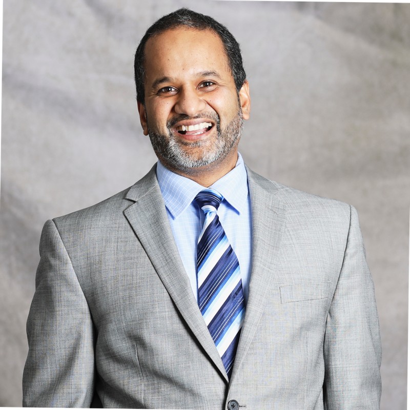 Smiling person with a beard wearing a light gray suit and a blue-striped tie against a gray background.
