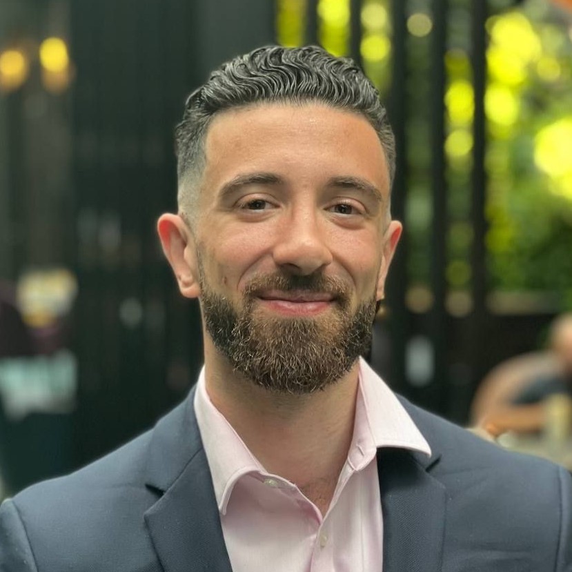 A man with short, curly dark hair and a trimmed beard smiles at the camera. He is wearing a light pink shirt and a dark blazer, standing outdoors with greenery and blurred lights in the background.