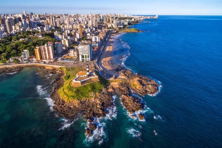 Aerial view of a coastal city with numerous buildings and high-rises along the shoreline. A prominent lighthouse on a rocky peninsula juts into the ocean, surrounded by vibrant blue water. The sky is clear, and the coastline extends to the horizon.