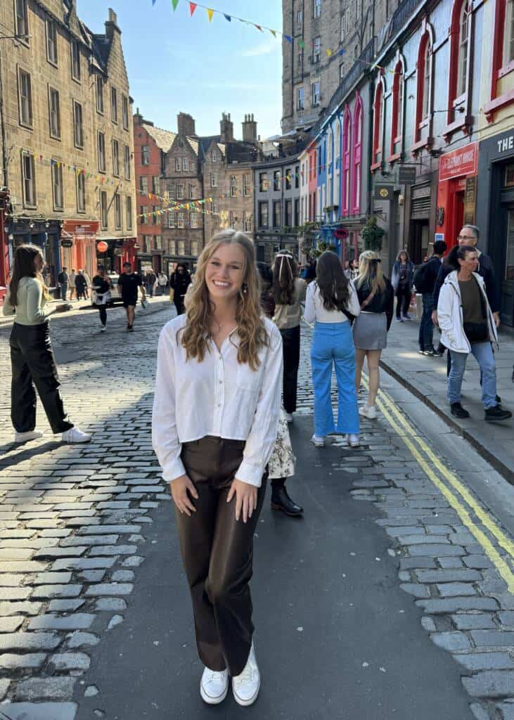 A young woman with long, wavy hair stands smiling on a cobblestone street. She wears a white shirt and dark pants. Behind her, people walk and chat, and the buildings lining the street are colorful, with strings of flags hanging between them.