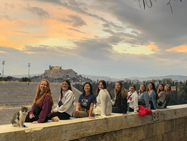 A group of nine people sitting on a stone wall, with a cat nearby. In the background, the Acropolis of Athens is visible under a cloudy sky at sunset. The atmosphere is relaxed and scenic.