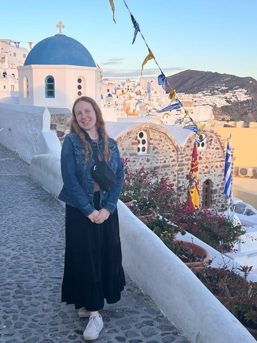 A person with long hair stands smiling on a stone pathway with traditional white buildings and a blue-domed church in the background. Colorful flags hang above, and the sky is clear. The setting is reminiscent of a Mediterranean town.