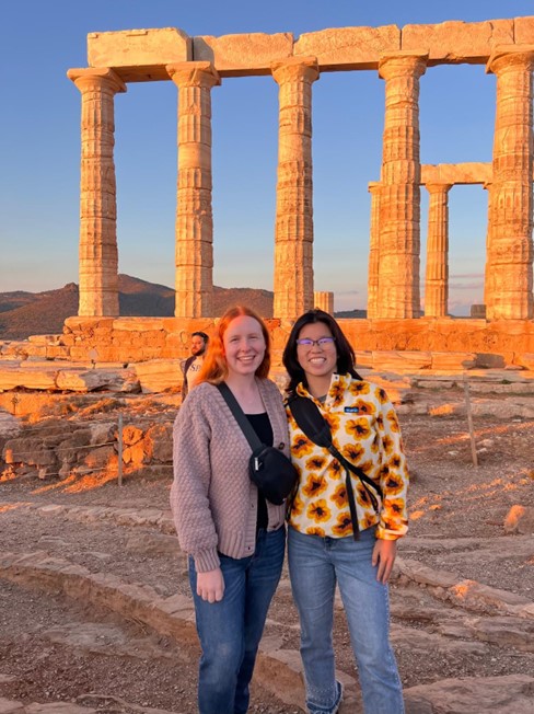 Two people smiling and posing in front of ancient Greek ruins at sunset, with tall marble columns in the background. One is wearing a pink cardigan, and the other has a sunflower-patterned jacket.