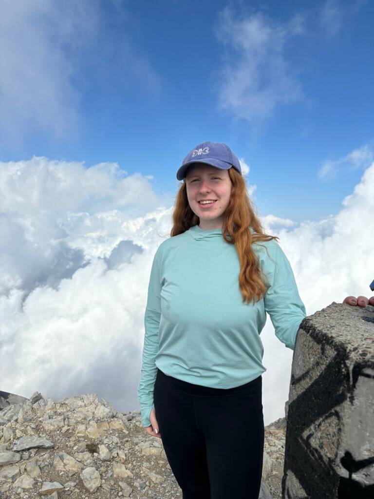 A person with long red hair, wearing a light blue long-sleeve shirt and blue cap, stands on a rocky mountain summit. They are smiling with a background of clouds and blue sky.
