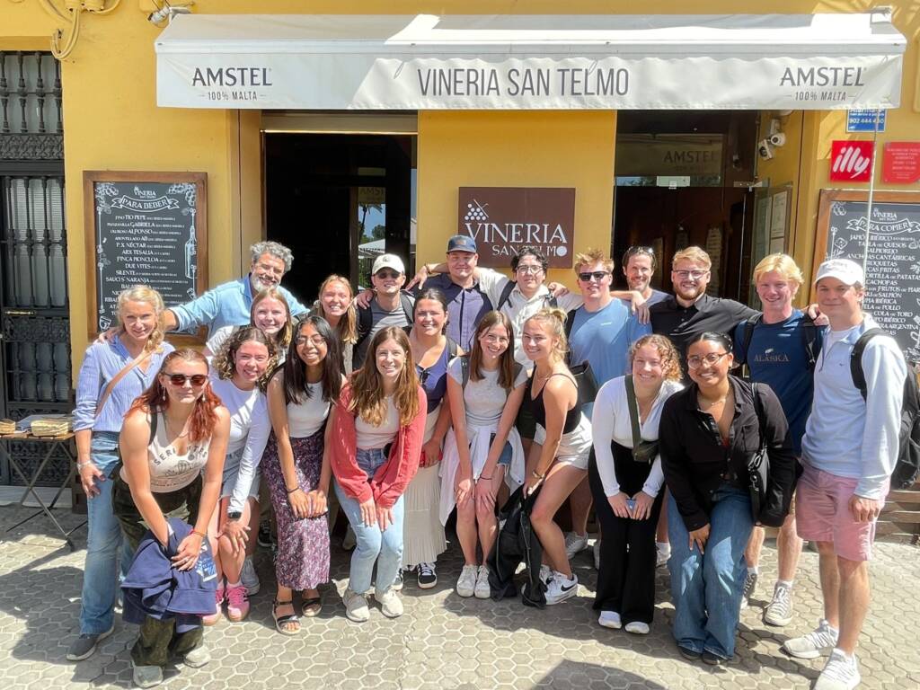 A group of smiling people gathered in front of Vineria San Telmo, a yellow building with signs for Amstel and illy coffee. Some are kneeling in front, and others are standing behind them, all posing together on a sunny day.