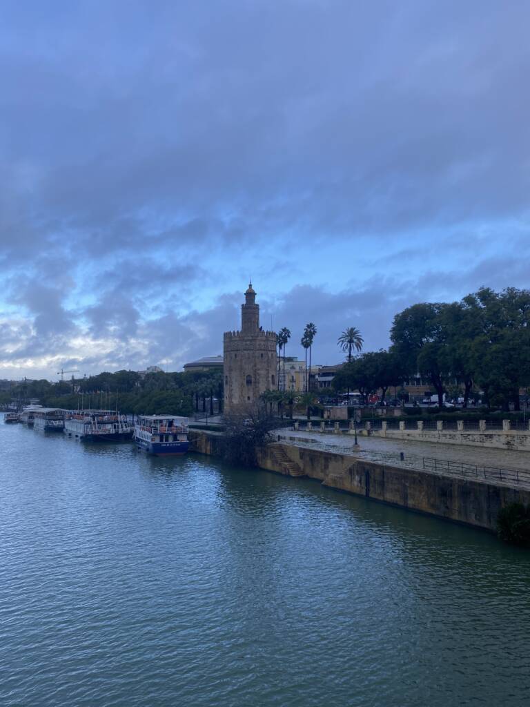A riverside view featuring a tall, historic tower surrounded by trees. Boats are docked along the water, and a cloudy sky looms overhead, casting a moody atmosphere. People are walking along the stone promenade.