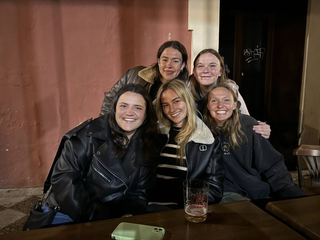 Five people sit closely together at a table, smiling warmly. Four wear jackets with fur collars, while one wears a striped top. A drink is on the table, and a wall with graffiti is in the background.
