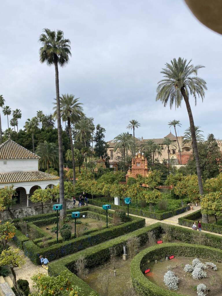 A lush garden with neatly trimmed hedges and tall palm trees under a cloudy sky. In the background, there are historic buildings with tiled roofs and a red brick structure. Pathways intersect through the garden, and a few visitors stroll around.