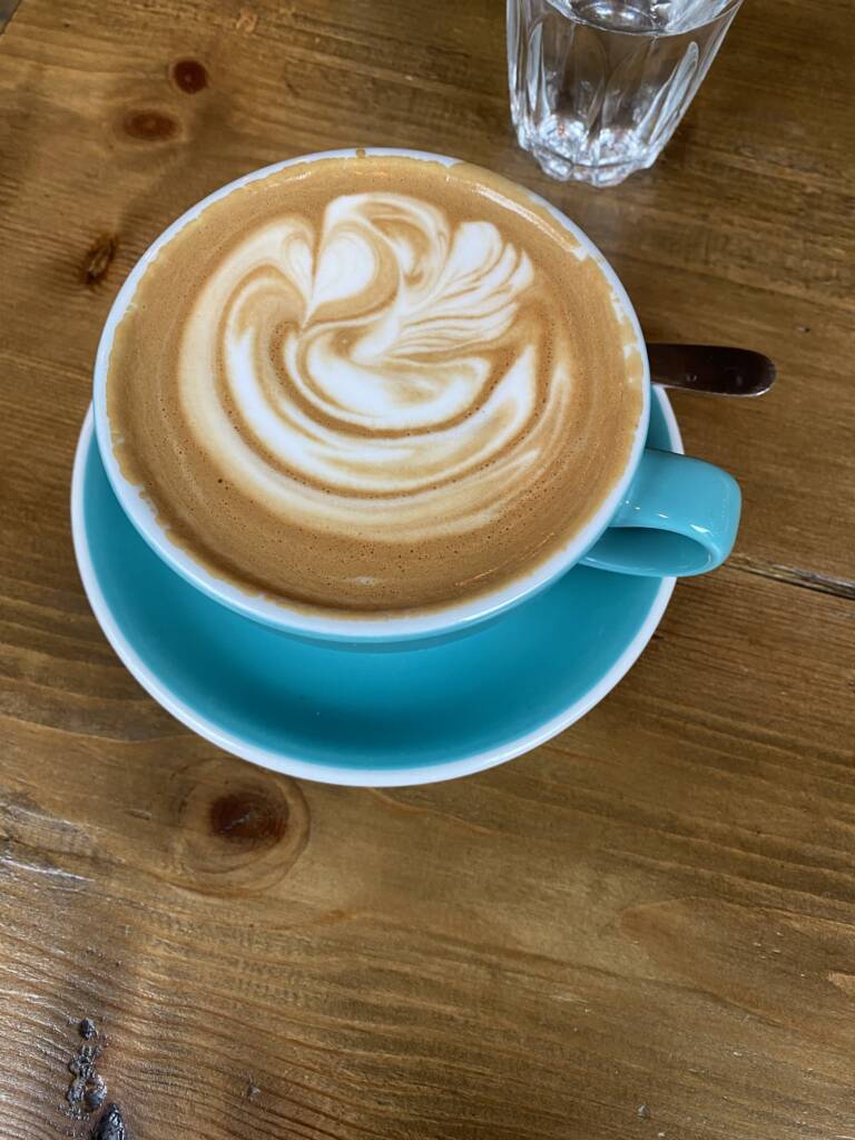 A latte in a teal cup with intricate foam art on top, set on a matching saucer on a wooden table. A small, clear glass of water is in the background.