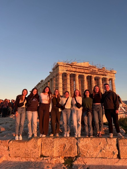 A group of people stand on a stone ledge at sunset in front of the Parthenon in Athens. They are lined up and smiling, with the ancient structure bathed in warm light behind them. Scaffolding is visible on parts of the Parthenon.
