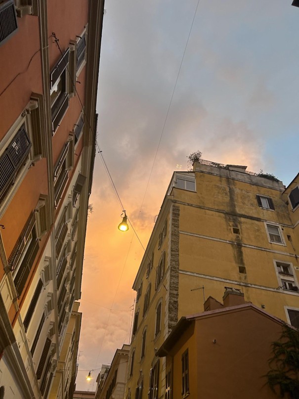 View of buildings from a narrow street at dusk, with a streetlamp glowing against a sky filled with warm, pastel-colored clouds. The buildings are in shades of orange and yellow, with some balconies and plants visible.