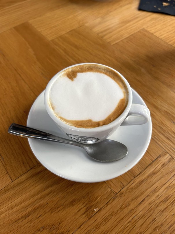 A small white cup of cappuccino with frothy milk sits on a matching saucer. A metal teaspoon is placed on the saucer. The background shows a wooden table surface with a herringbone pattern.