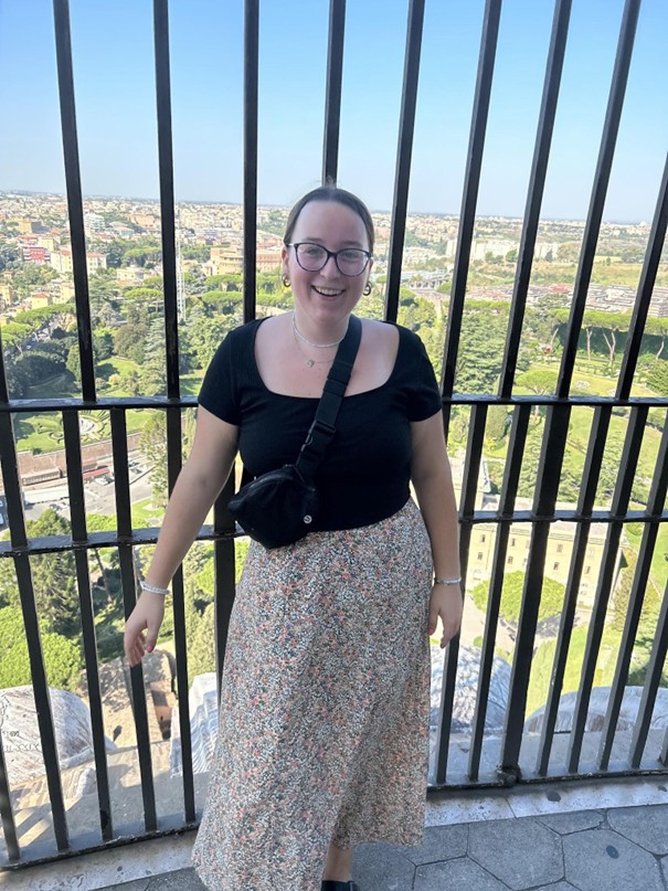 A person with glasses and a black top smiles while standing in front of a railing with a scenic view of a city and greenery in the background. They are wearing a floral skirt and a black crossbody bag.