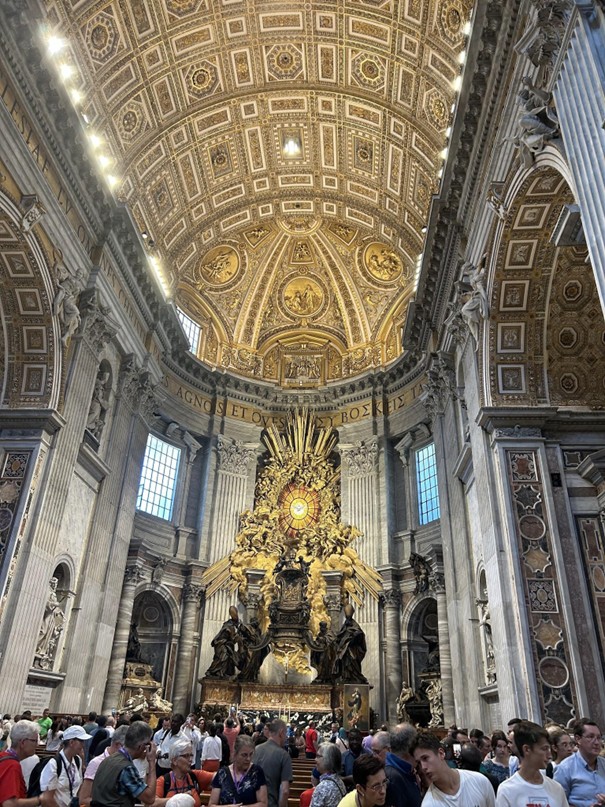 People gather inside a grand cathedral with an ornately decorated dome and intricate architectural details. The ceiling features elaborate gold designs and statues, with a large sculptural centerpiece near the altar.