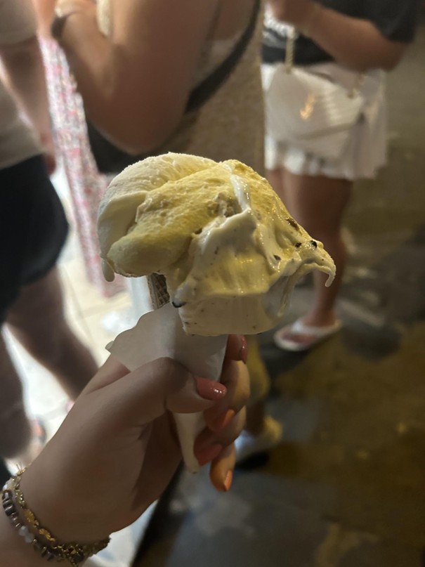 A person is holding a melting ice cream cone with multiple flavors in a dimly lit area. The background is blurred, showing a small group of people and a sidewalk.