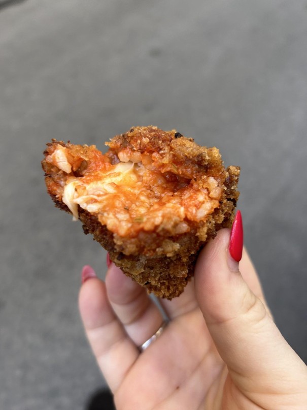 A hand with red nails holds a partially eaten fried risotto ball. The inside reveals a mix of rice and a red sauce, likely tomato-based, with a bit of visible cheese. The background is a blurred gray surface.