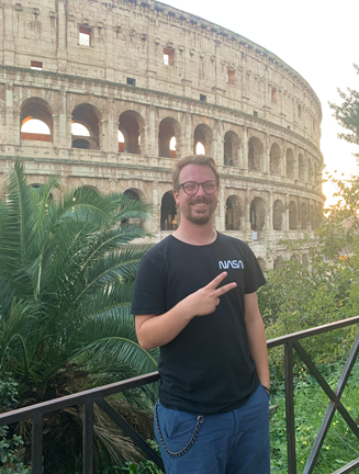A person wearing glasses and a black NASA t-shirt stands smiling and holding up a peace sign, in front of the Colosseum in Rome. There are green plants in the foreground.