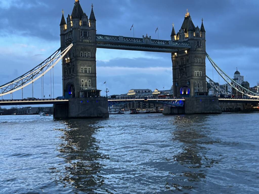Tower Bridge in London illuminated at dusk, with lights reflecting on the Thames River. The sky is cloudy, creating a moody atmosphere. Buildings are visible in the background.