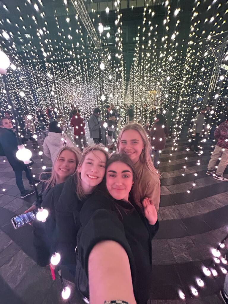 Four friends take a selfie in a dazzling installation of hanging lights. The scene is vibrant, with strands of glowing bulbs creating a festive atmosphere. Other people are visible in the background, enjoying the display.