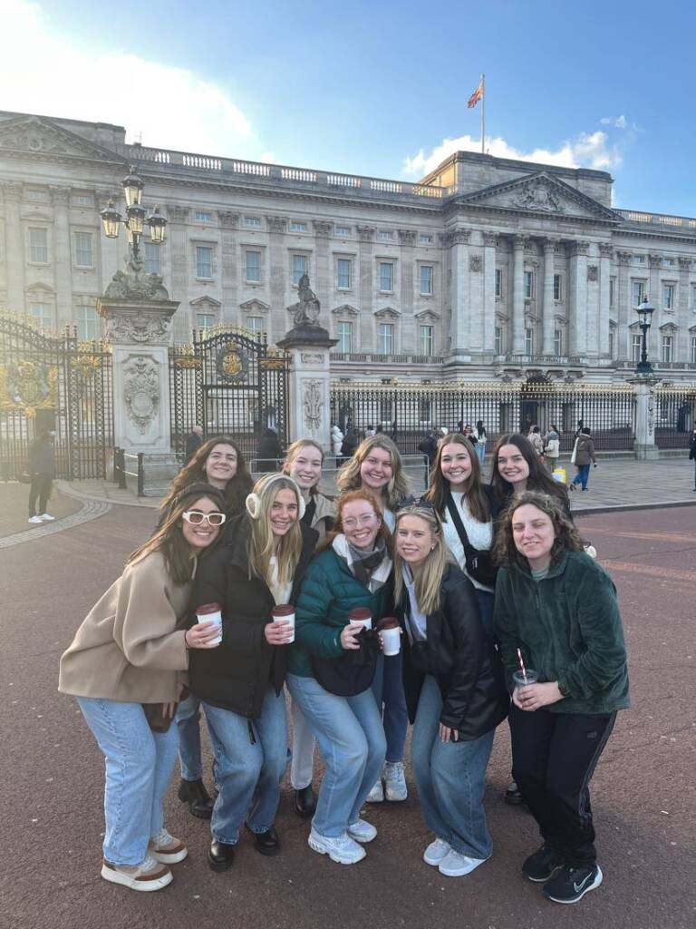 A group of twelve people pose happily with drinks in front of Buckingham Palace. The sky is clear and sunny. They are dressed in casual winter clothing, and the palace's ornate gates are visible in the background.