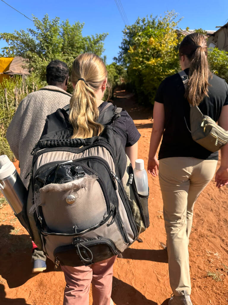 Three people walking on a dirt path surrounded by greenery. The person in the foreground wears a large backpack with water bottles and has hair in a ponytail. The sun casts shadows as they walk away from the camera.