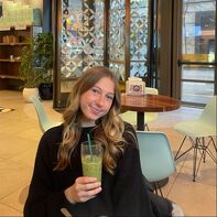 A person with long hair sits at a round table in a cafe, smiling and holding a green drink with a straw. The cafe features modern decor, with light-colored chairs and geometric patterns on the windows.