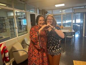 Two people stand close together in an office setting. The person on the left wears a red patterned dress, while the person on the right wears glasses, a black top, and brown pants. They are forming a heart shape with their hands and smiling at the camera.
