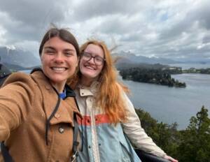 Two people smiling for a selfie in front of a scenic background. They are dressed in jackets, suggesting cool weather. Behind them, there's a large, calm body of water surrounded by trees and distant mountains under a cloudy sky.