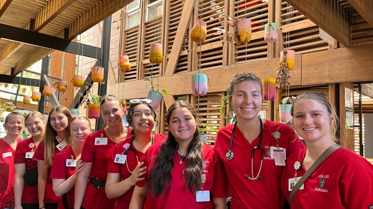 A group of smiling nursing students in red uniforms pose together indoors in front of a wall decorated with colorful hanging planters and wooden slats.