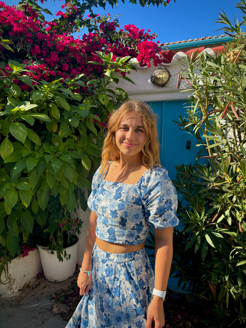A woman in a blue floral two-piece outfit stands in bright sunlight, surrounded by lush green plants and vibrant pink flowers, with a blue door and white wall in the background.