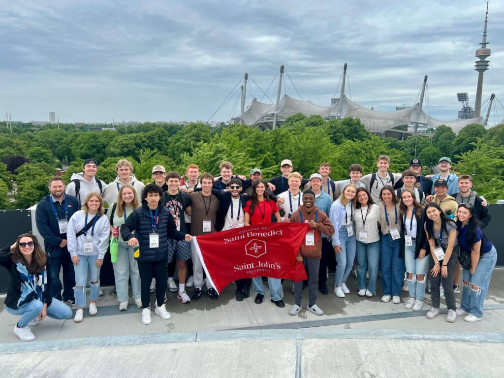 A group of young adults pose together outdoors, smiling in front of lush green trees and a modern stadium structure. Several in front hold a red “Saint Benedict Saint John’s” banner. The sky is cloudy.